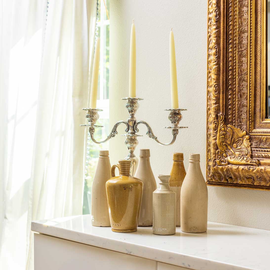 Decorative vases and candles on a windowsill with a gold-framed mirror in the background.