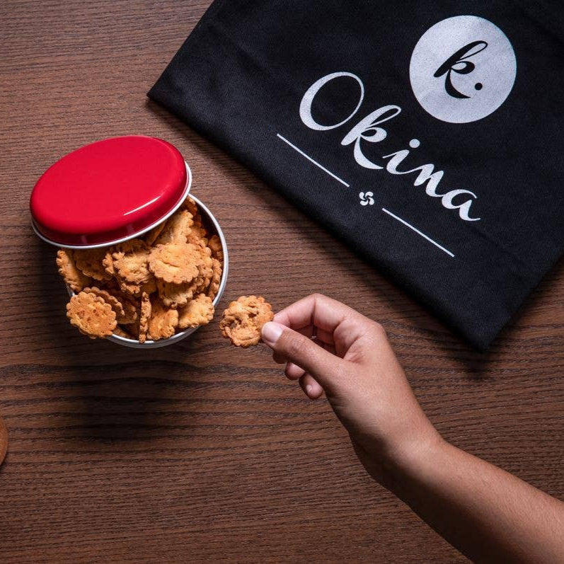 Container of snacks with a red lid, wooden spoon with spice, and hand reaching for a snack on a wooden surface with 'Okina' branding.