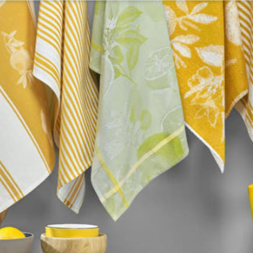 Set of yellow and white patterned towels hanging on a gray wall with a bowl and lemons in the foreground.