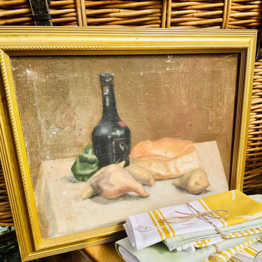 Framed still life painting with a bottle and bread on a tablecloth.