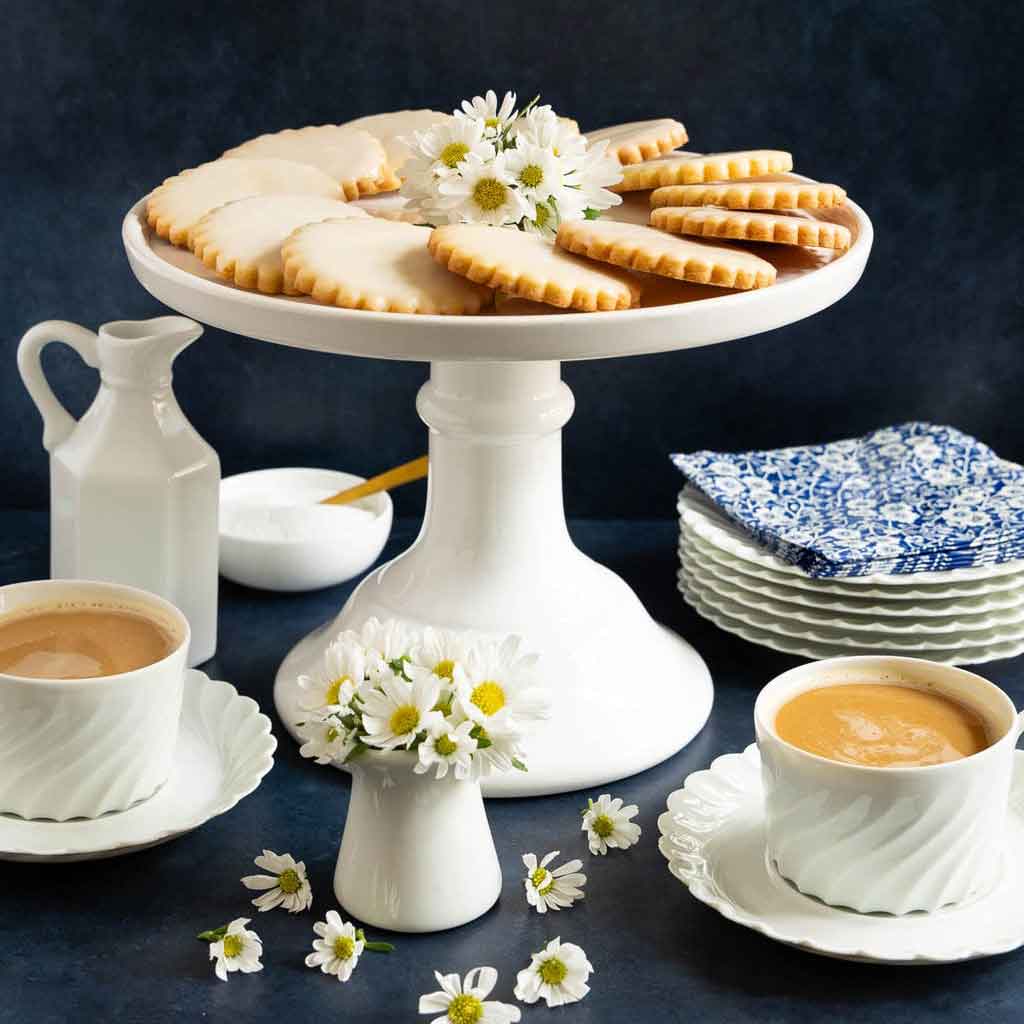 White cake stand with cookies, cups of coffee, and floral decorations on a dark surface.