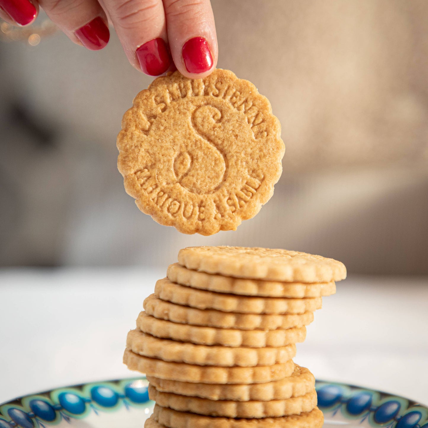 Hand holding a cookie above a stack of cookies on a decorative plate with a blurred background