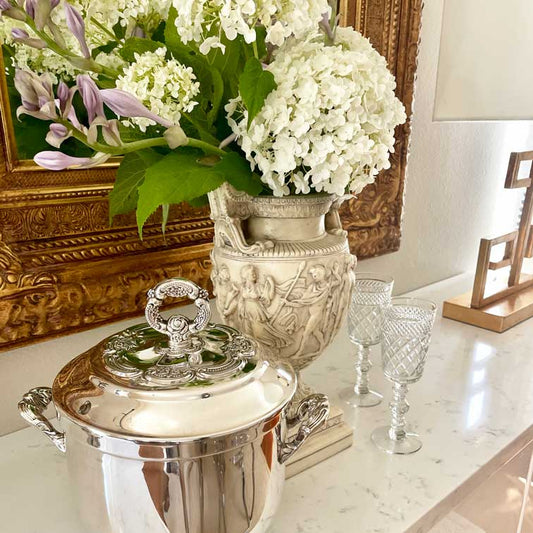 Decorative silver container, crystal glasses, and floral arrangement on a marble surface with a gold frame in the background.