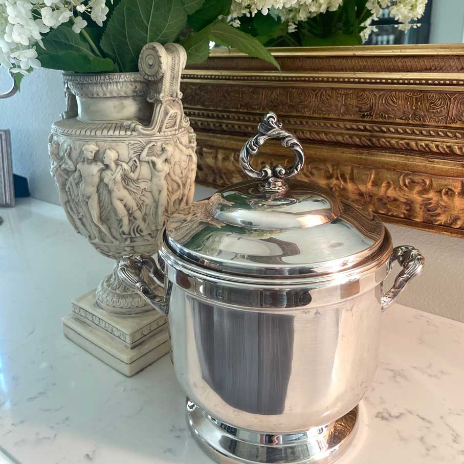 Silver ice bucket with decorative lid on a marble surface with a gold-framed mirror and decorative urn in the background.