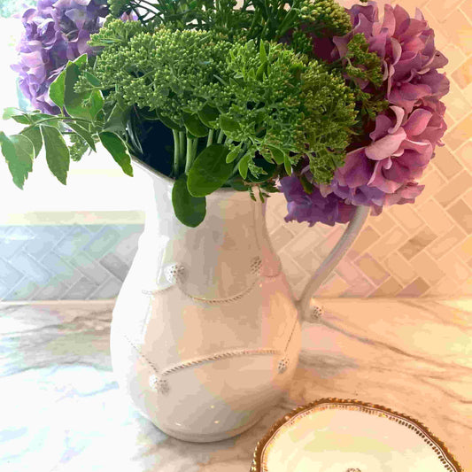 White vase with greenery and purple flowers on a light surface with a patterned wall in the background