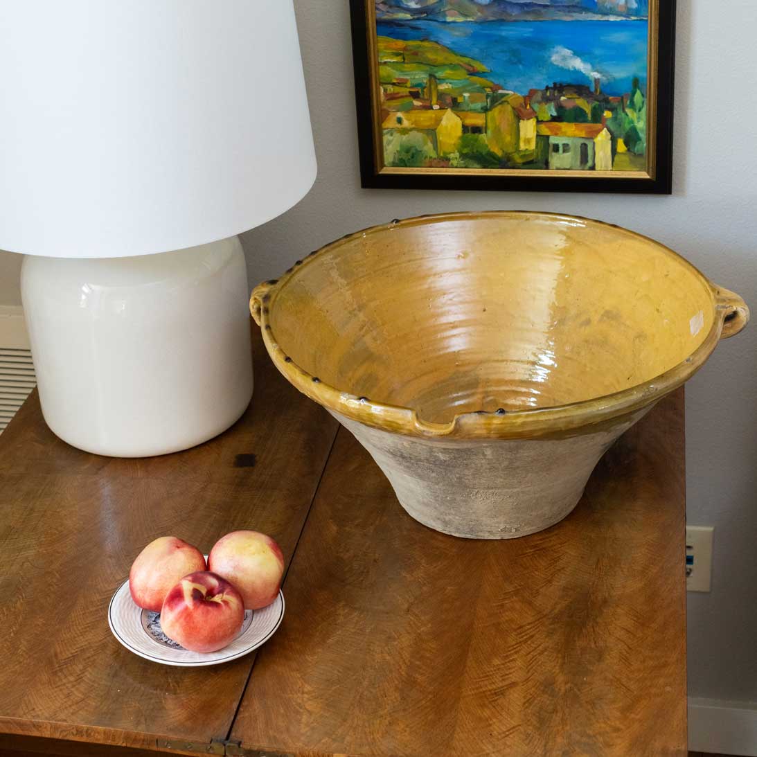 Yellow ceramic bowl on a wooden table with peaches and a lamp in the background.
