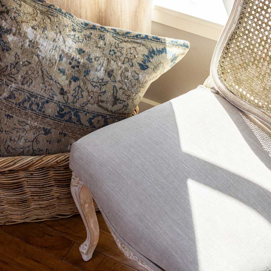 Decorative pillow on a wicker basket next to a white cushioned chair with woven backrest.